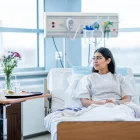 A young woman of Middle Eastern decent sits up in her hospital bed after surgery.  She is wearing a gown and is tucked warmly under a blanket and she looks down contemplatively.