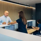 A senior male patient fills out a medical form while standing at the reception desk with the help of a friendly female medical worker
